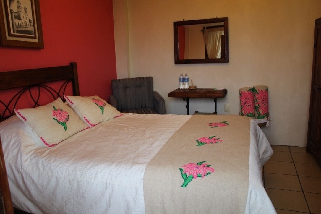 Een net opgemaakt bed met een bloemenbedekker, een stoel, een tafeltje met een fles water, en een spiegel aan de muur.