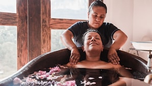 Una persona recibiendo un masaje en la espalda en una tina de madera llena de agua y flores rosas.