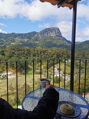A balcony with a clear view of a mountainous landscape, a table with a cup and a plate, and a person holding a cup.