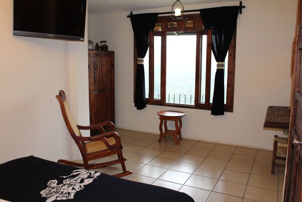 Una habitación con una cama, un sillón mecedora, una mesita, una ventana con cortinas y un televisor en la pared.