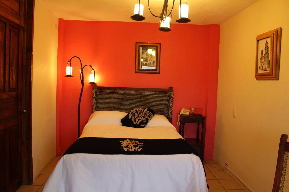 Ein Hotelzimmer mit roter Wand, einem Bett mit schwarz-weiß gestreifter Bettdecke, zwei Wandlampen, einem gerahmten Bild an der Wand und einer Holztür.