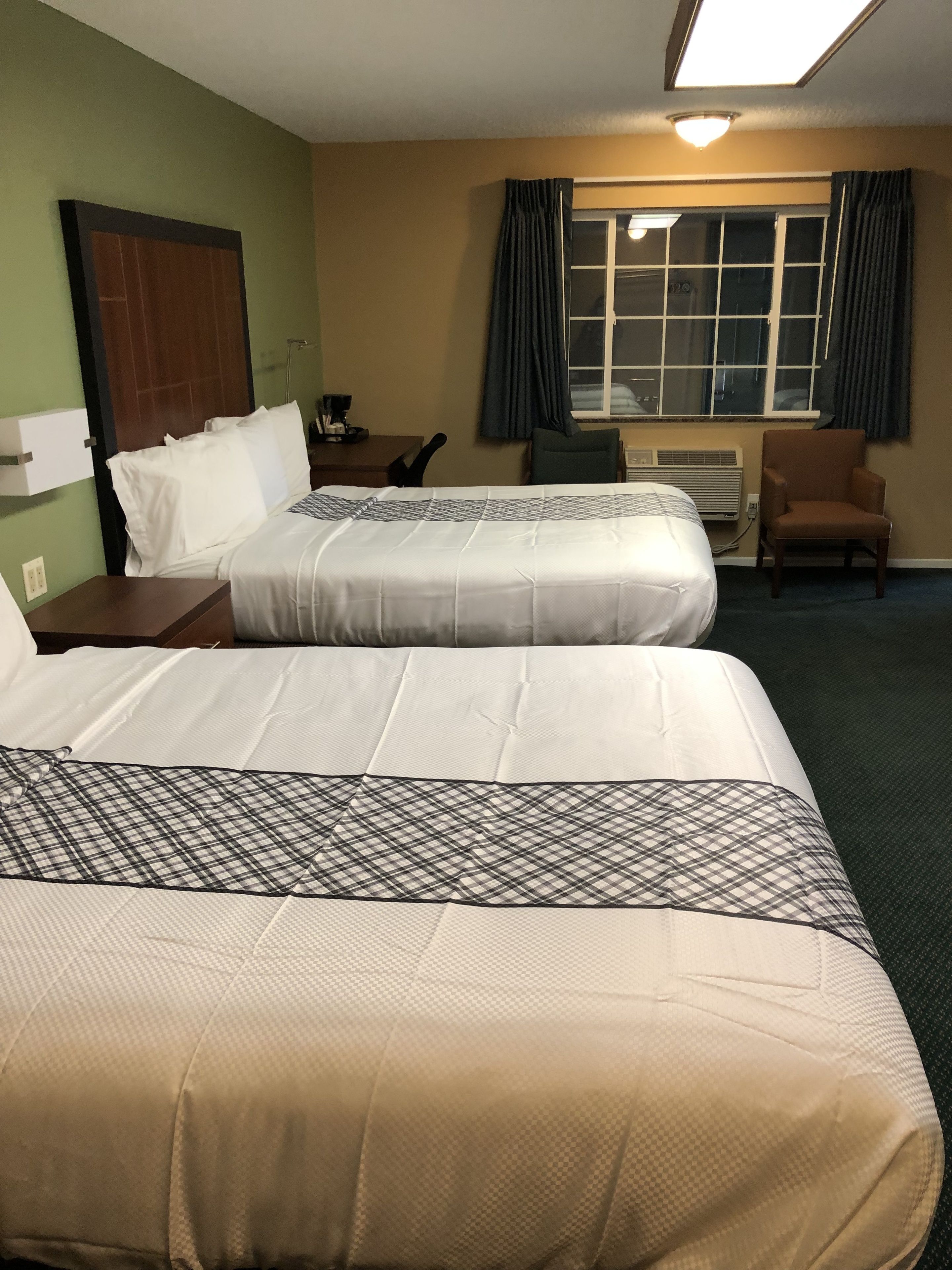 Habitación de hotel con dos camas, un escritorio, una silla, un ventanal con cortinas y un equipo de aire acondicionado.