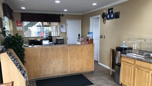 A reception area with a wooden counter, a computer, and a coffee machine. There is a door, a plant, and a black mat on the floor. - Knight's Inn Motel (Grants Pass)