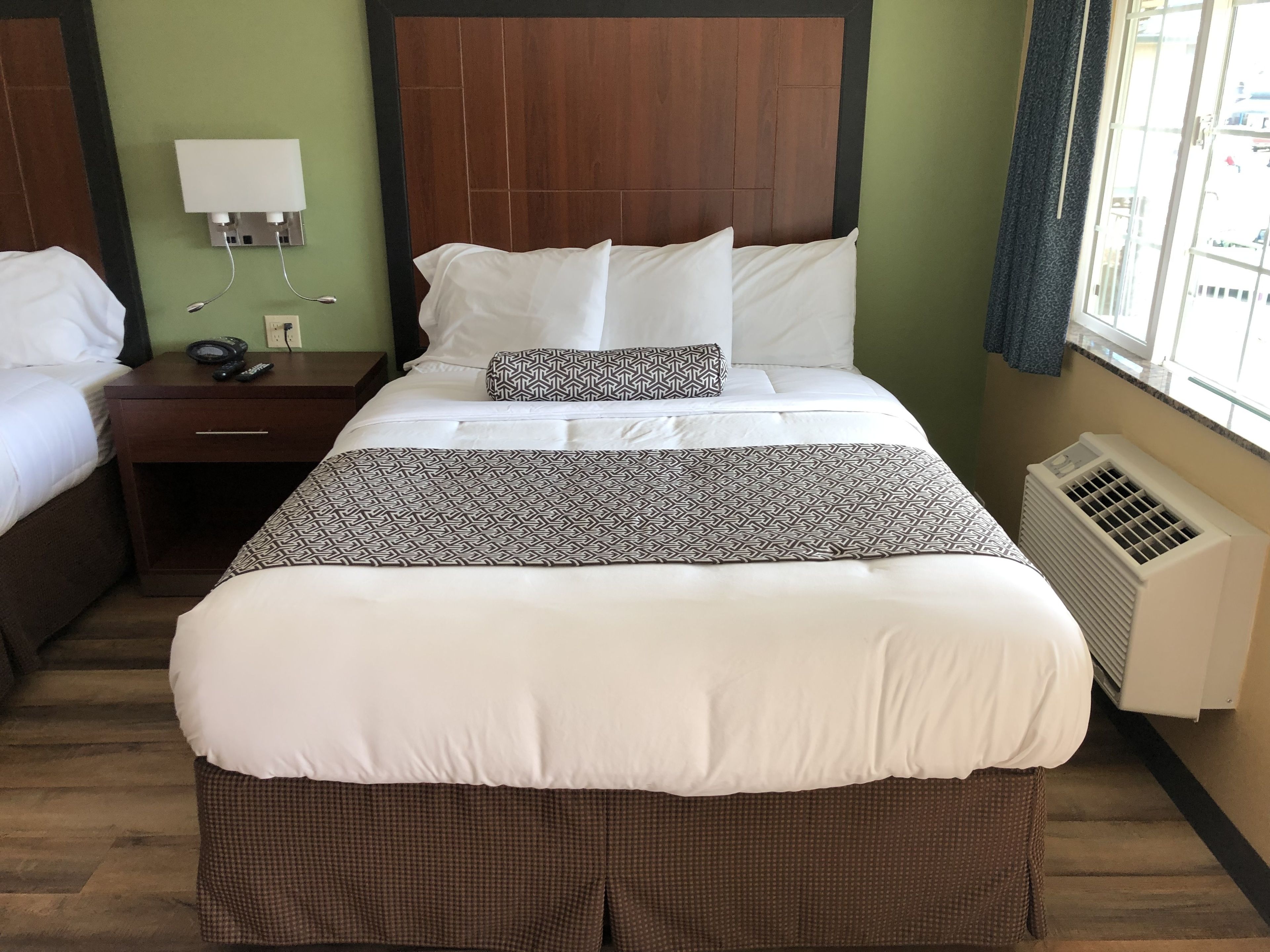 A hotel room with a large bed, a bedside table with a phone, a wall socket, and a window with curtains.