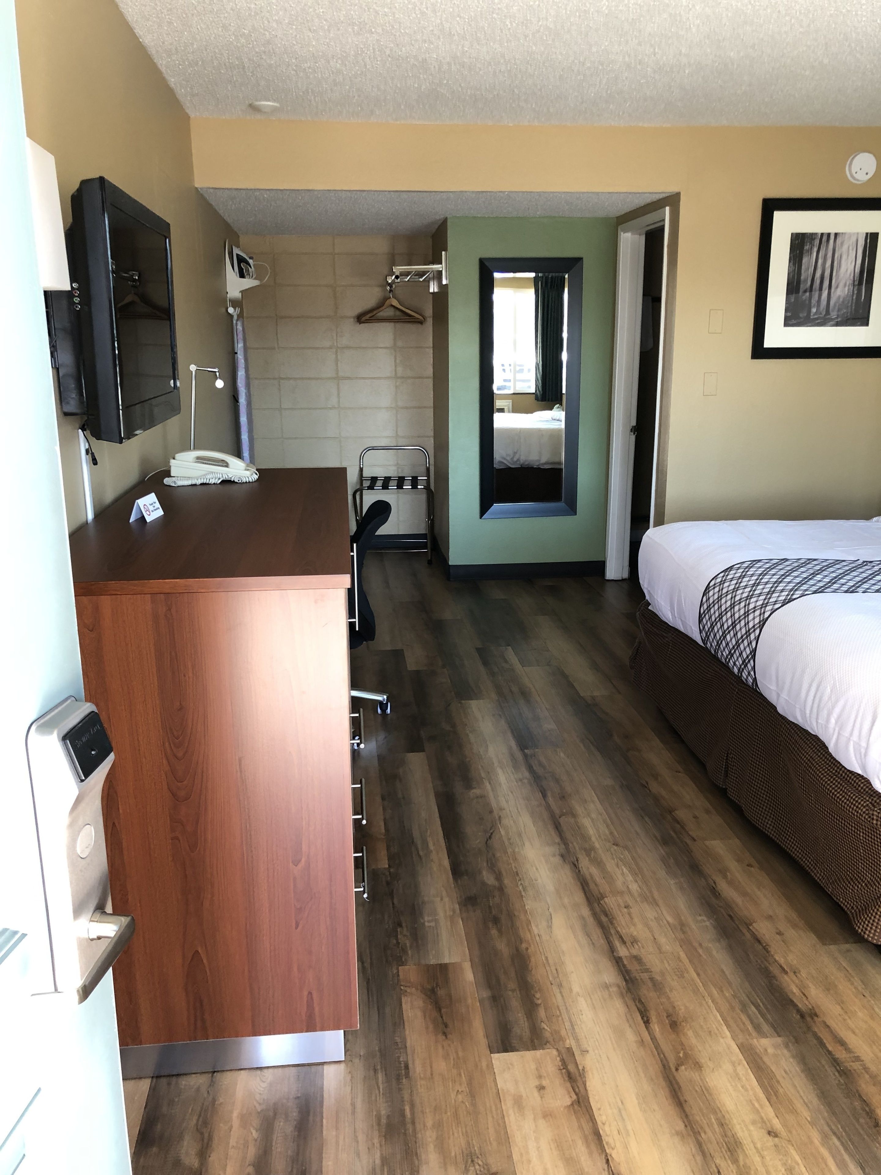 A hotel room with a wooden desk, a bed, a mirror, and a hanging coat rack.