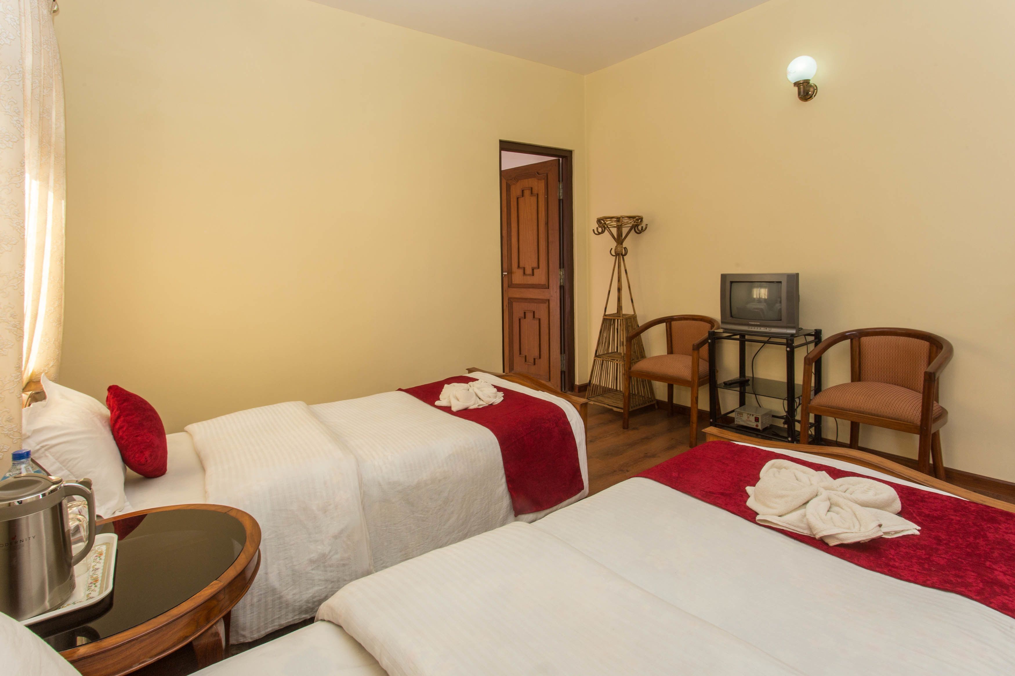 A hotel room with two beds, a small table with a kettle, a chair, a TV, and a lamp.