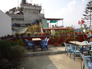 A rooftop terrace with blue plastic chairs and tables, surrounded by potted plants and a view of a building with a red roof. - Hotel Earth House (Kathmandu)