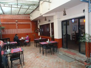 An outdoor dining area with brick walls, tables with red placemats, and hanging bells. - Hotel Earth House (Kathmandu)