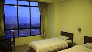 Ein Hotelzimmer mit zwei Betten, einem Schreibtisch und einem großen Fenster mit Blick auf die Stadt.