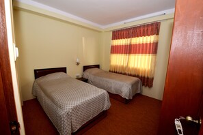 A room with two single beds, a wooden headboard, a nightstand, a wall-mounted lamp, and a window with striped curtains. - Kirtipur Hillside Hotel and Resort (Kirtipur)