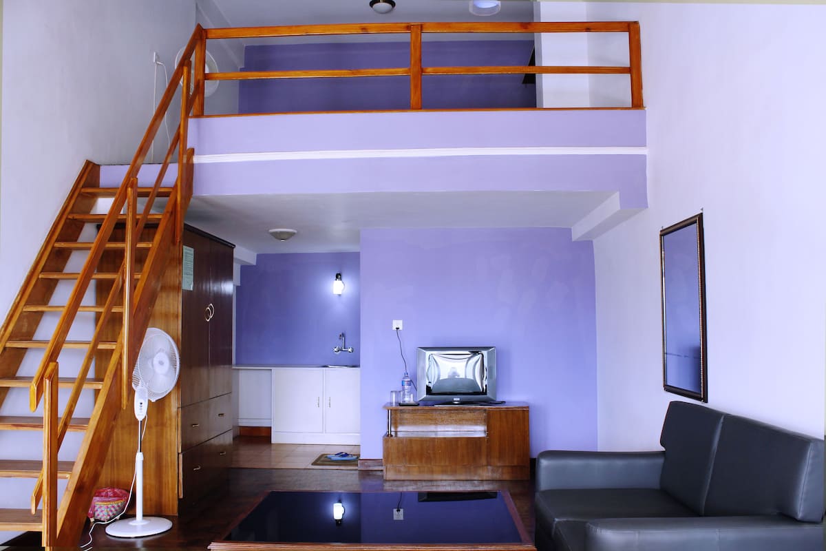 A multi-level living space with a wooden staircase, a flat-screen TV on a wooden stand, a black leather sofa, and a small table.