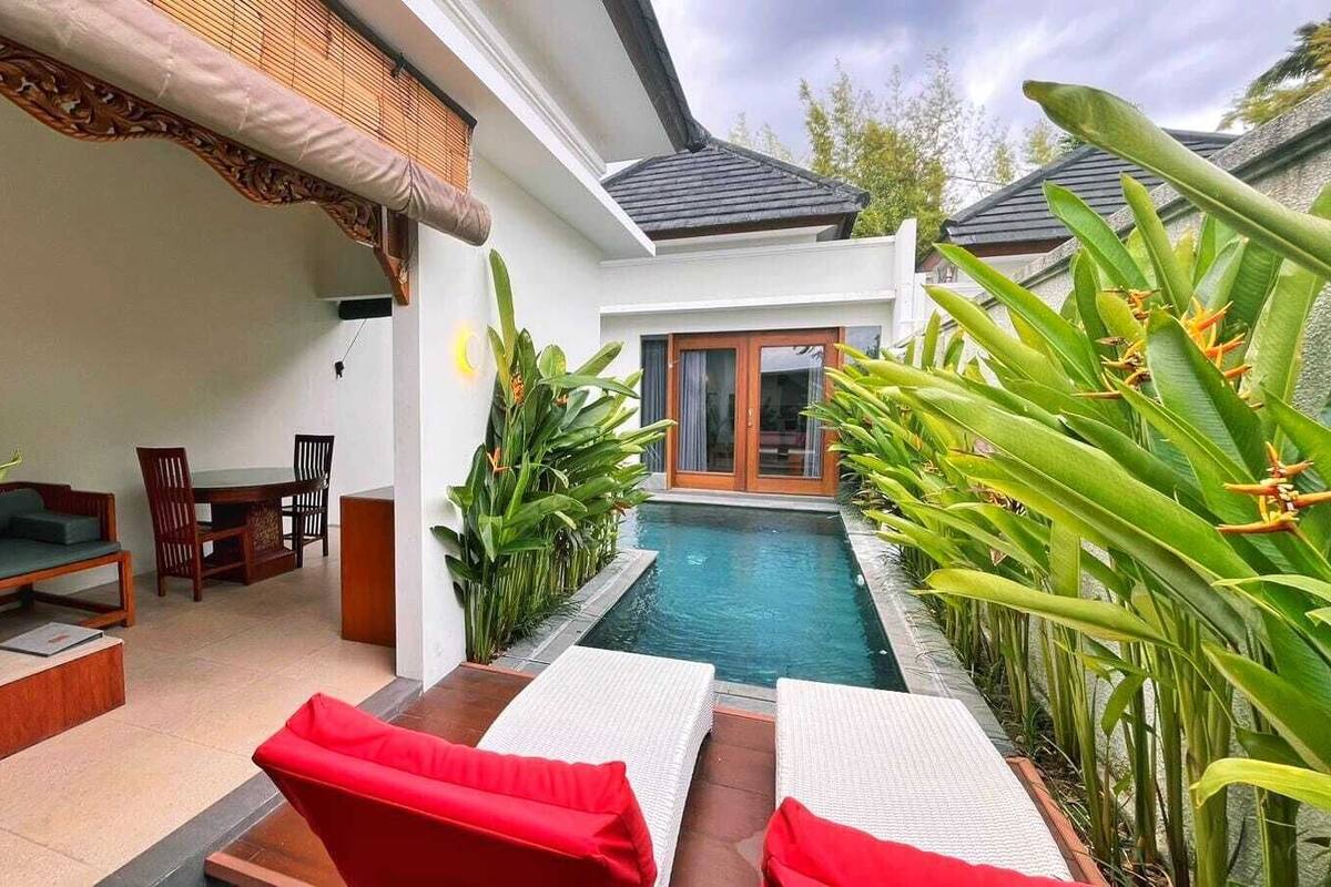 Un área junto a la piscina con sillones de descanso, un conjunto de comedor y plantas tropicales.