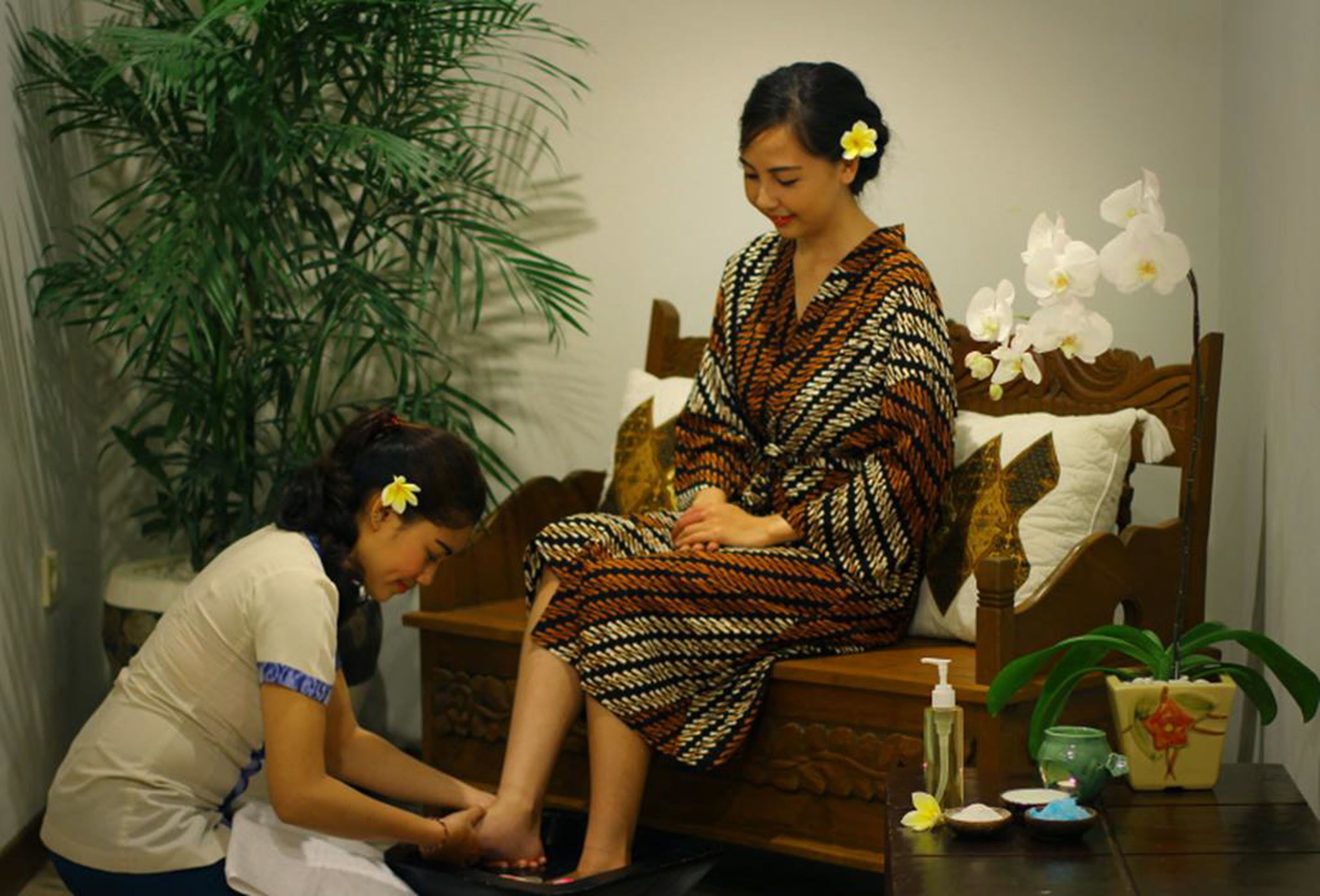 A woman receiving a foot massage in a room with a wooden bed, a large green plant, and a small table with toiletries.