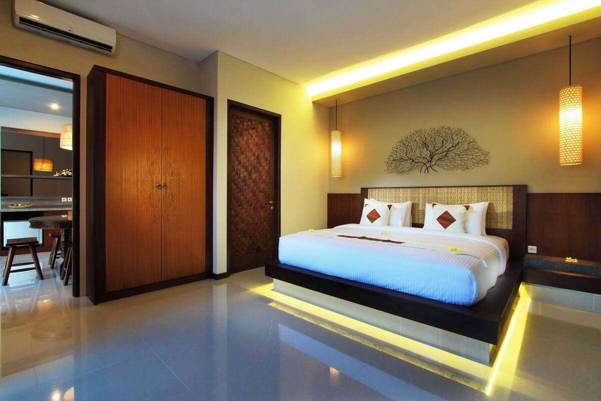 A modern hotel room with a large bed, a wooden wardrobe, a desk with chairs, and a wall-mounted light fixture.