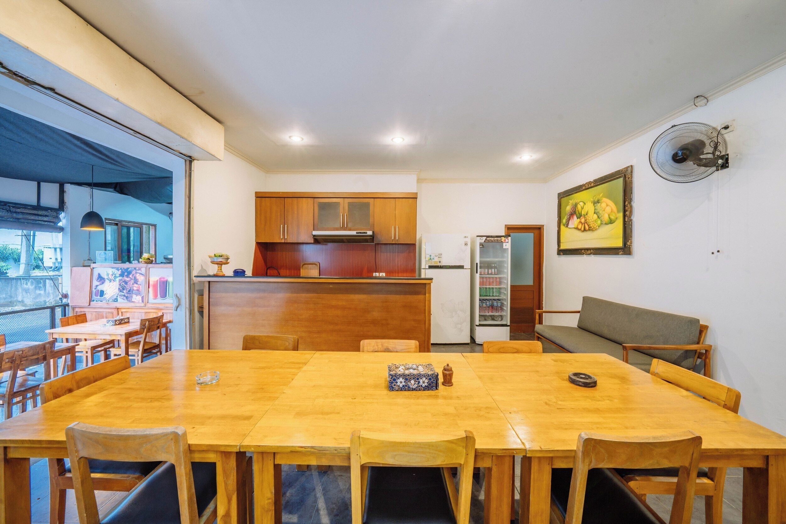 A dining area with a wooden table and chairs, a kitchen in the background, and a fan on the wall.