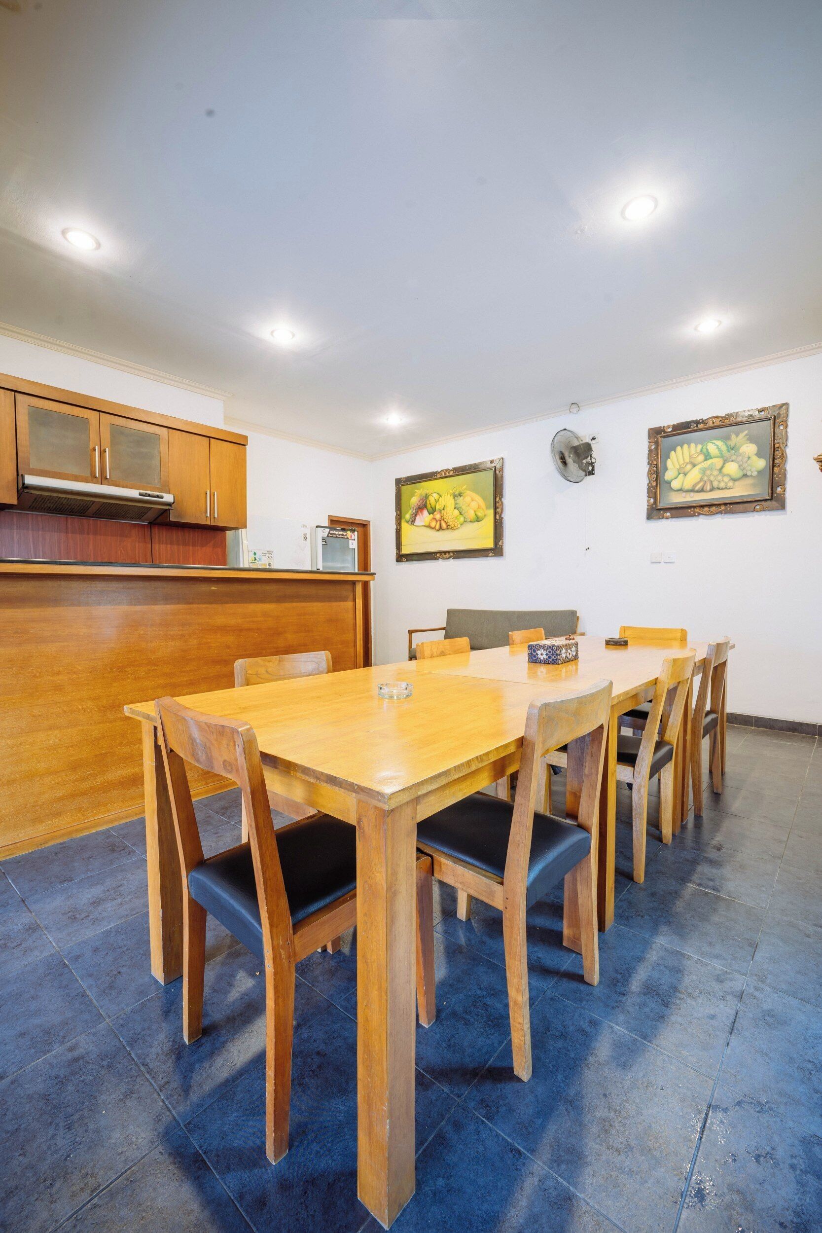 A dining area with a wooden table and chairs, a kitchen with wooden cabinets, and two framed paintings on the wall.