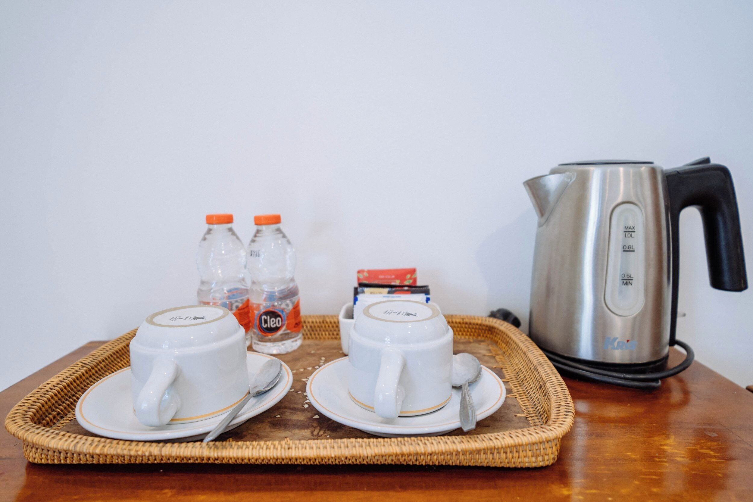 A tray with two white cups and saucers, two bottles of water, a box of tissues, and a silver electric kettle on a wooden table.