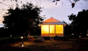 A small, single-story structure with a pitched roof, possibly a gazebo or a small pavilion, surrounded by trees and lit by lanterns.
