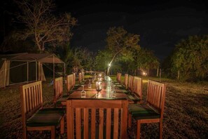 A long wooden table set for dining outdoors at night, surrounded by wooden chairs, with a tent and trees in the background.