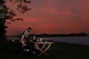 Un grupo de personas sentadas en una mesa de madera al aire libre al anochecer, con una masa de agua y árboles de fondo.