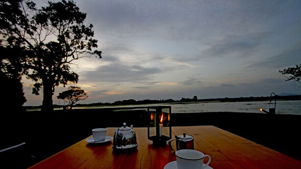 A wooden table set with a teapot, cups, and a lantern, overlooking a body of water at dusk. - The Yala Camping (Tissamaharama)