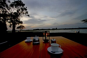 Una mesa de madera dispuesta con un calentador de agua, tazas y un farol, con vistas a una masa de agua al anochecer.