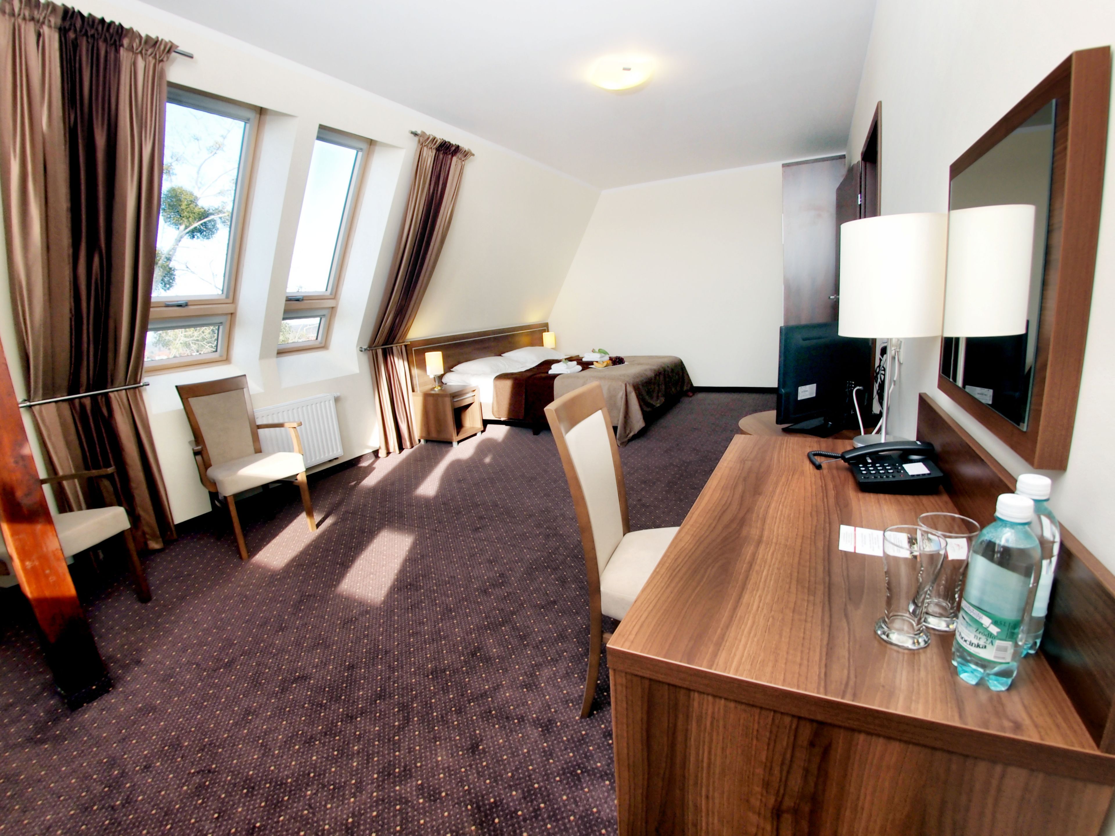 A hotel room with a bed, a desk with a phone and glasses, a chair, a TV, and a window with curtains.