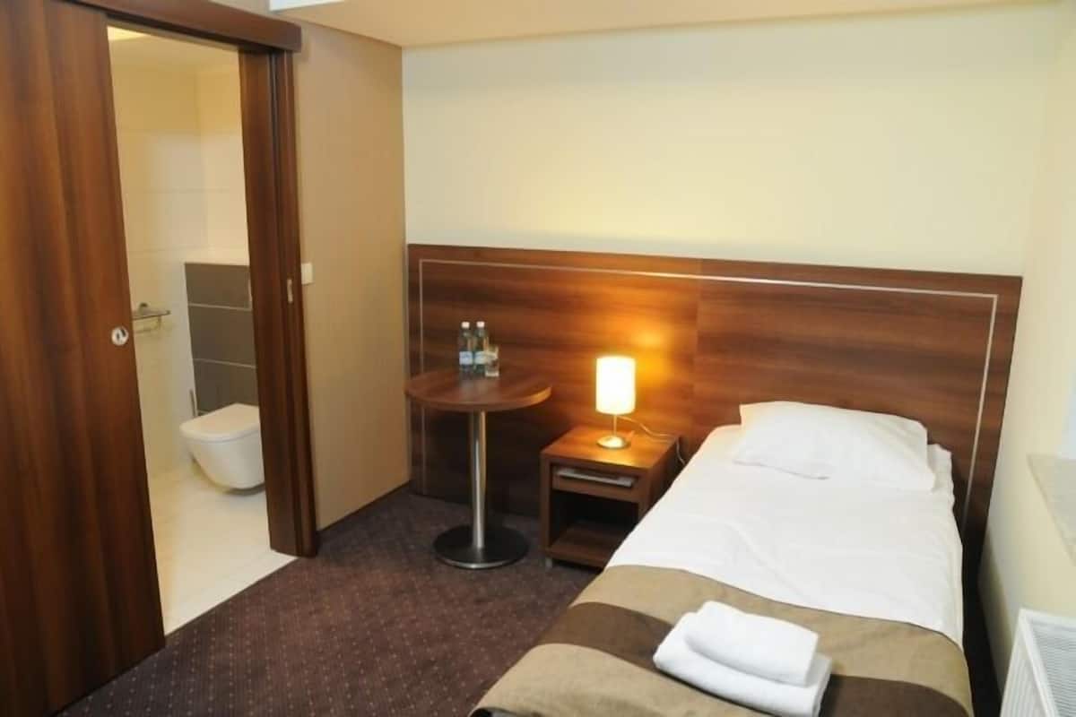 A hotel room with a bed, a bedside table with a lamp, a small round table, and a bathroom visible through an open door.