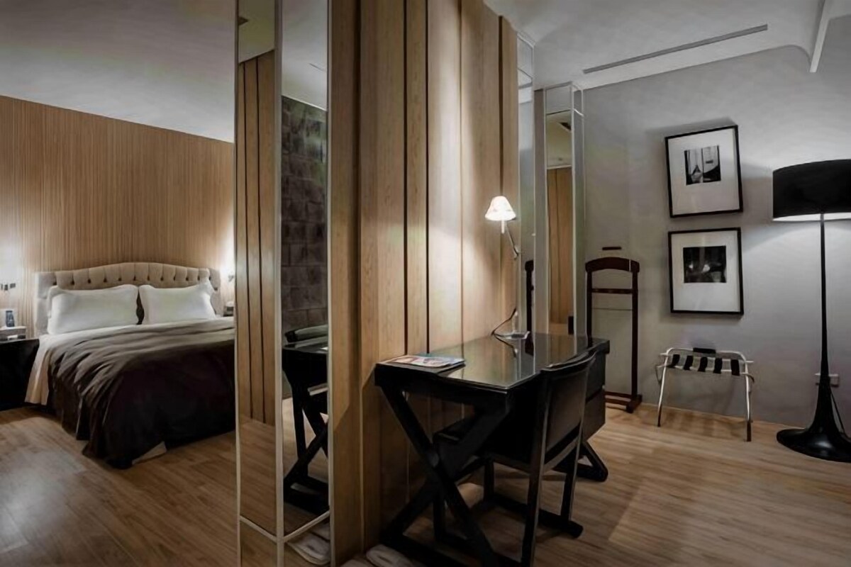 A hotel room with a bed, desk, chair, lamp, and framed pictures on the wall.