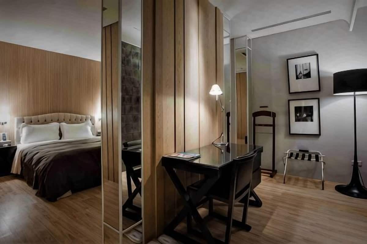 A hotel room with a bed, desk, chair, lamp, and framed pictures on the wall.