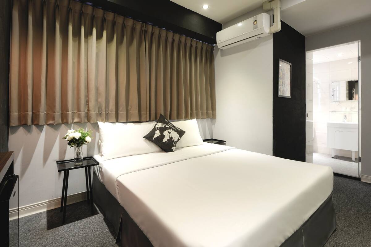 Quarto de hotel moderno com uma cama grande, uma mesa de cabeceira, um vaso com flores e um aparelho de ar condicionado.
