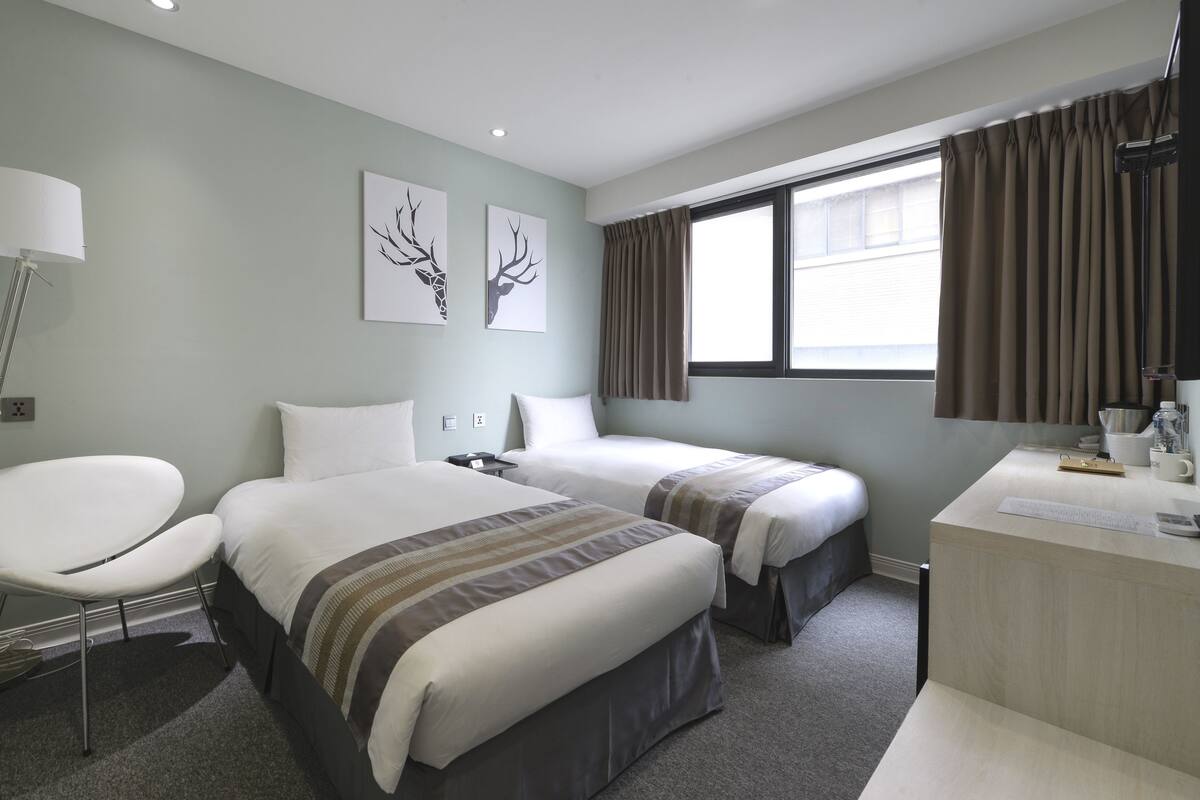 Quarto de hotel com duas camas, uma escrivaninha, uma cadeira, uma luminária e obras de arte na parede.