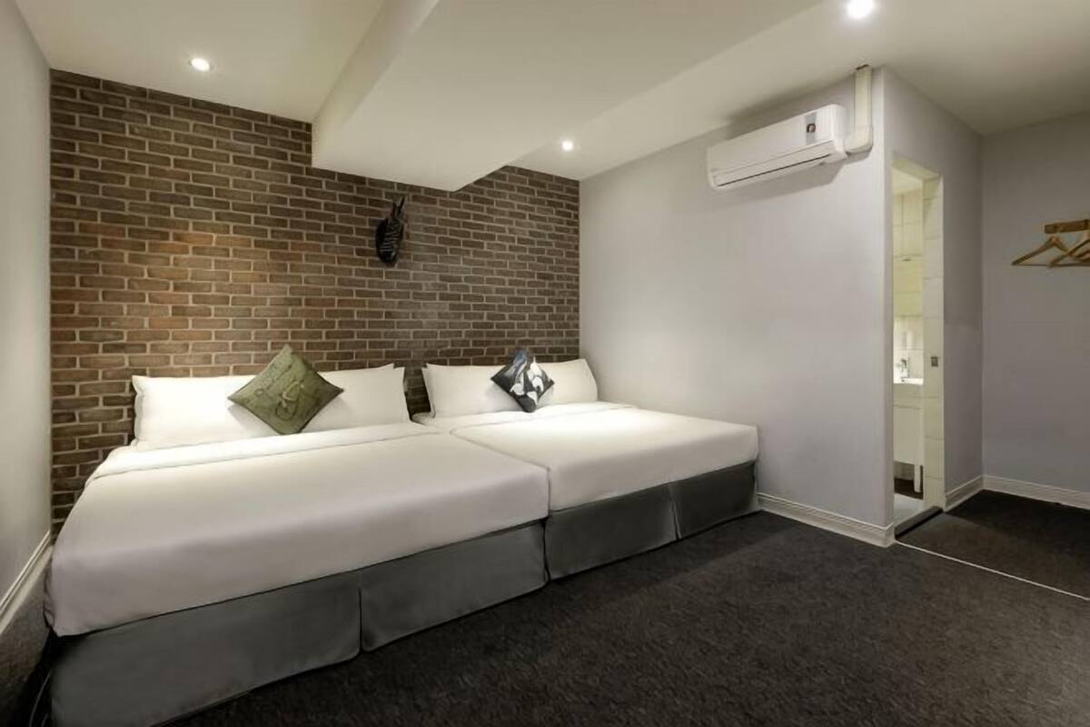 Quarto de hotel moderno com parede de destaque em tijolo, duas camas, carpete escuro e unidade de ar condicionado visível.