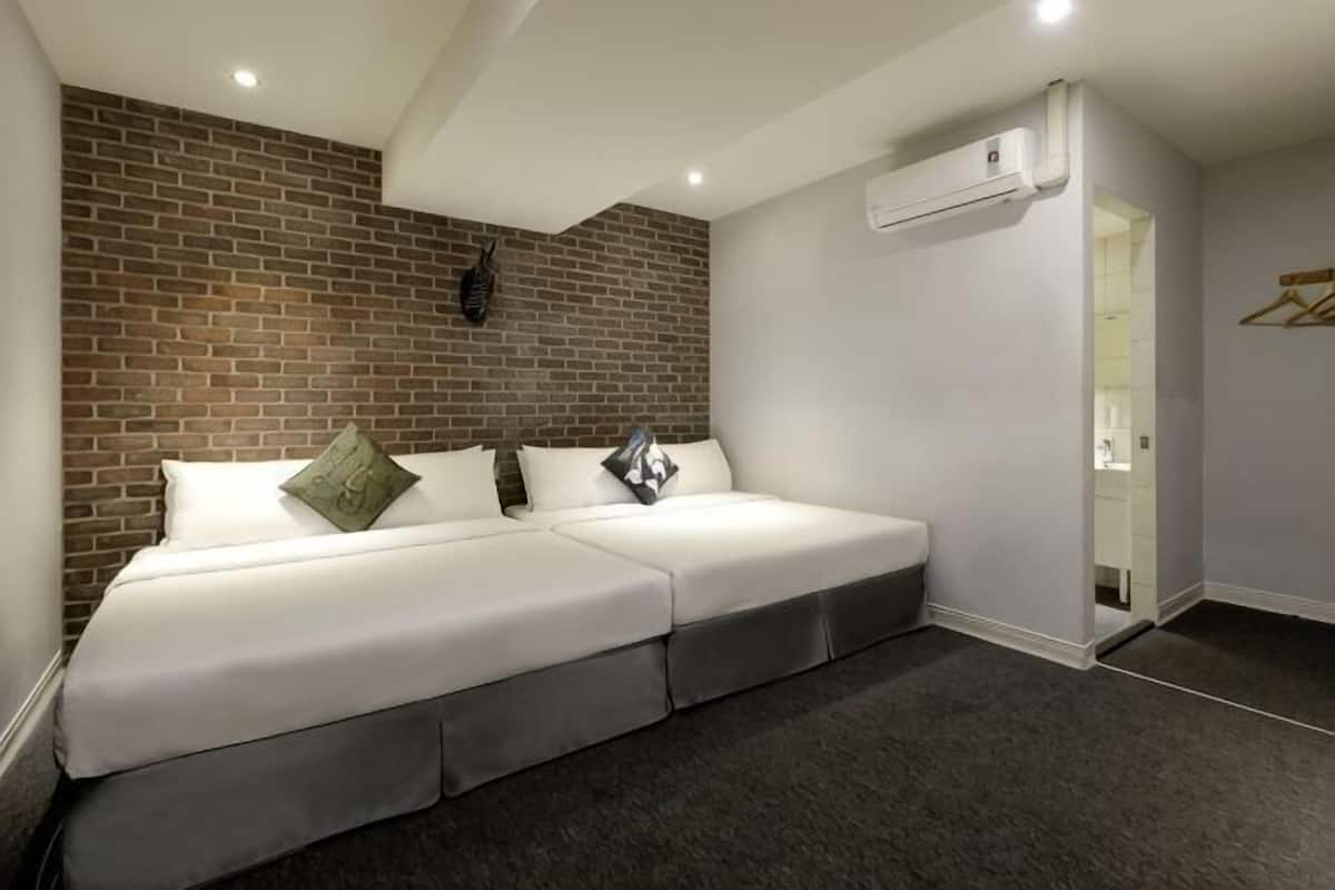 A modern hotel room with a brick accent wall, a two-bed setup, a dark carpet, and a visible air conditioning unit.