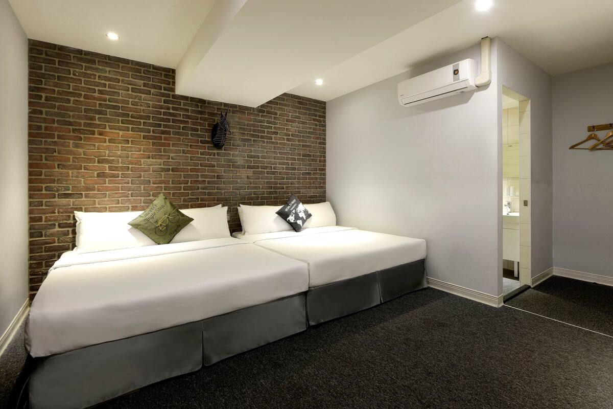 Quarto de hotel moderno com parede de destaque em tijolo, uma cama grande com roupa de cama branca e piso escuro coberto por carpete.