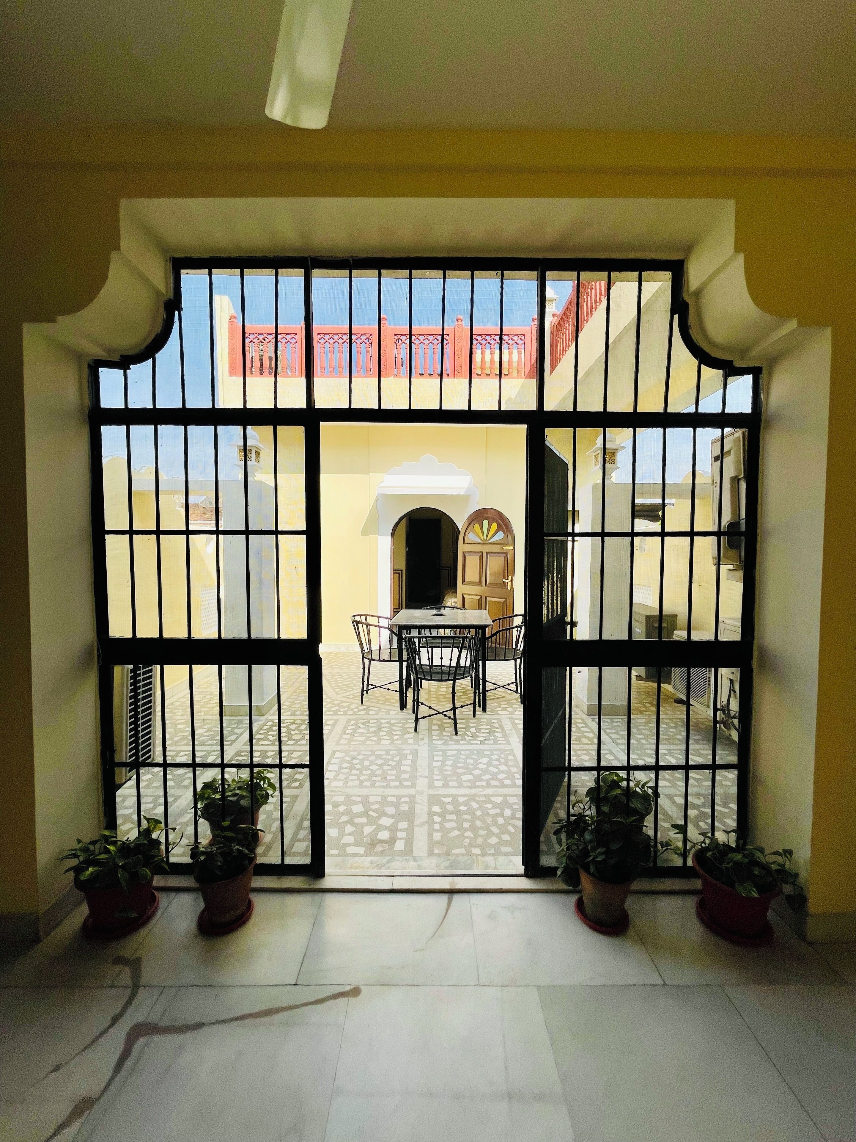 A view through a barred entrance to a courtyard with a tiled floor, a table, and chairs, and potted plants.