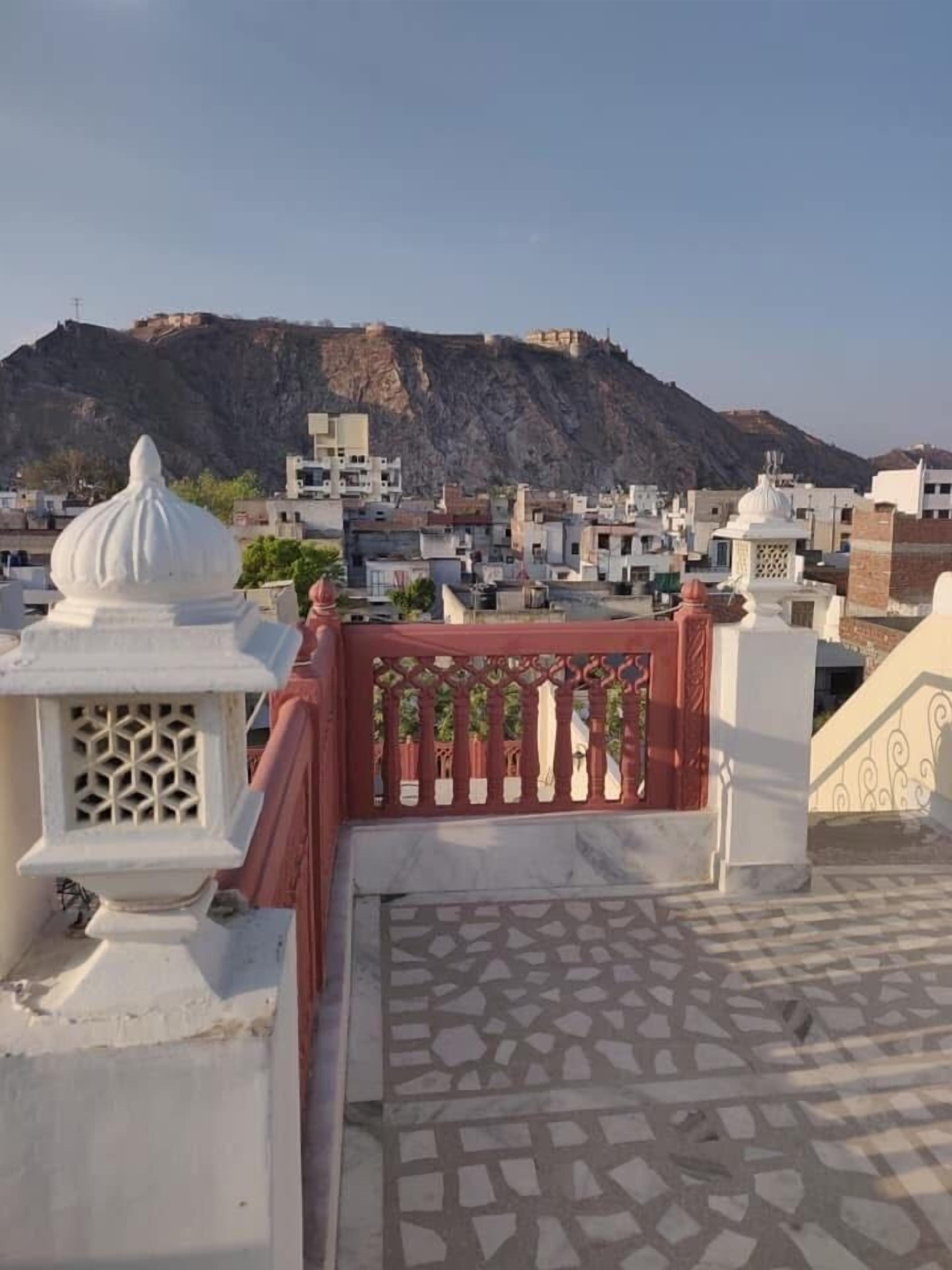 A rooftop terrace with a patterned floor, a red railing, and a view of a hill with buildings in the background.
