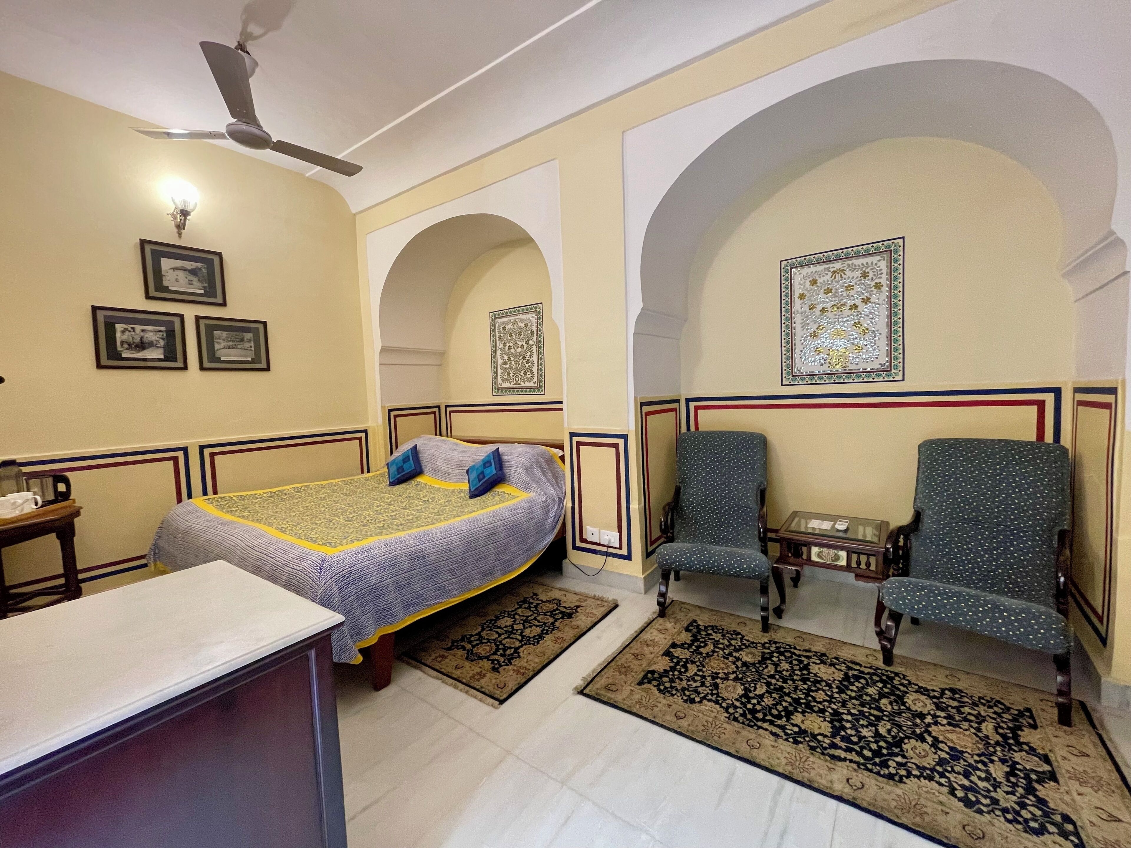 Une chambre d’hôtel avec un lit, deux fauteuils, un bureau et un ventilateur de plafond.