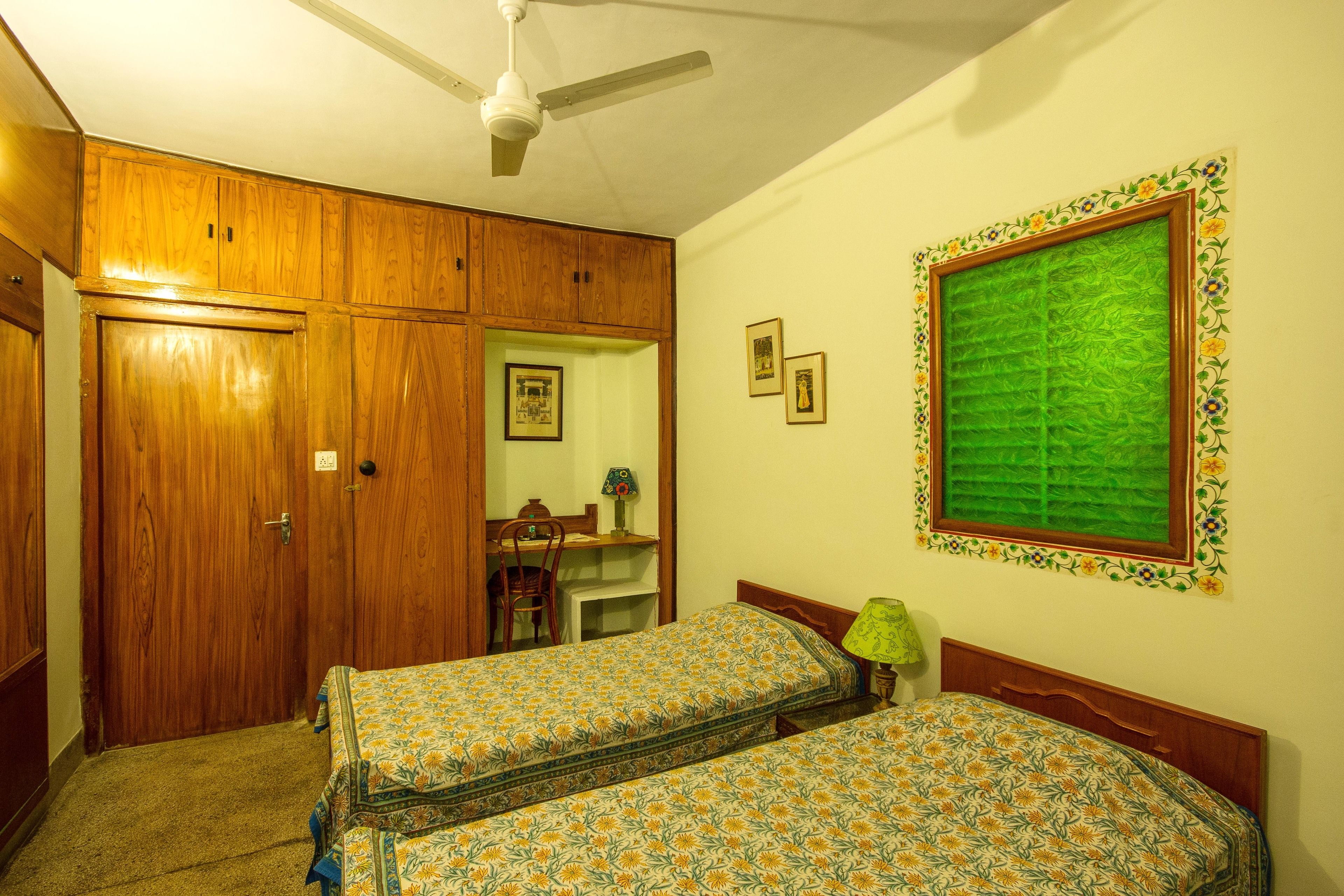Un dormitorio con dos camas, armario de madera, ventilador de techo, escritorio, silla y un cuadro en la pared.