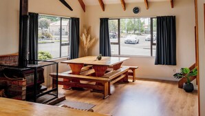 Amplia habitación con techo alto, vigas de madera, un ventanal con cortinas, una mesa de madera y una chimenea.