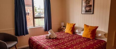 Une chambre d’hôtel avec un grand lit, deux oreillers, une chaise, une fenêtre avec des rideaux, une table de chevet avec une lampe et un tableau encadré au mur.