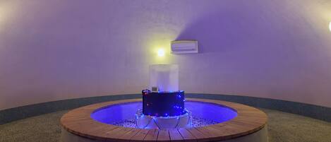 Une piscine ronde en bois, dont le centre est illuminé de lumière bleue, entourée d’un bord en béton, dans une pièce dont le mur est violet et qui est équipée d’un ventilateur de plafond.