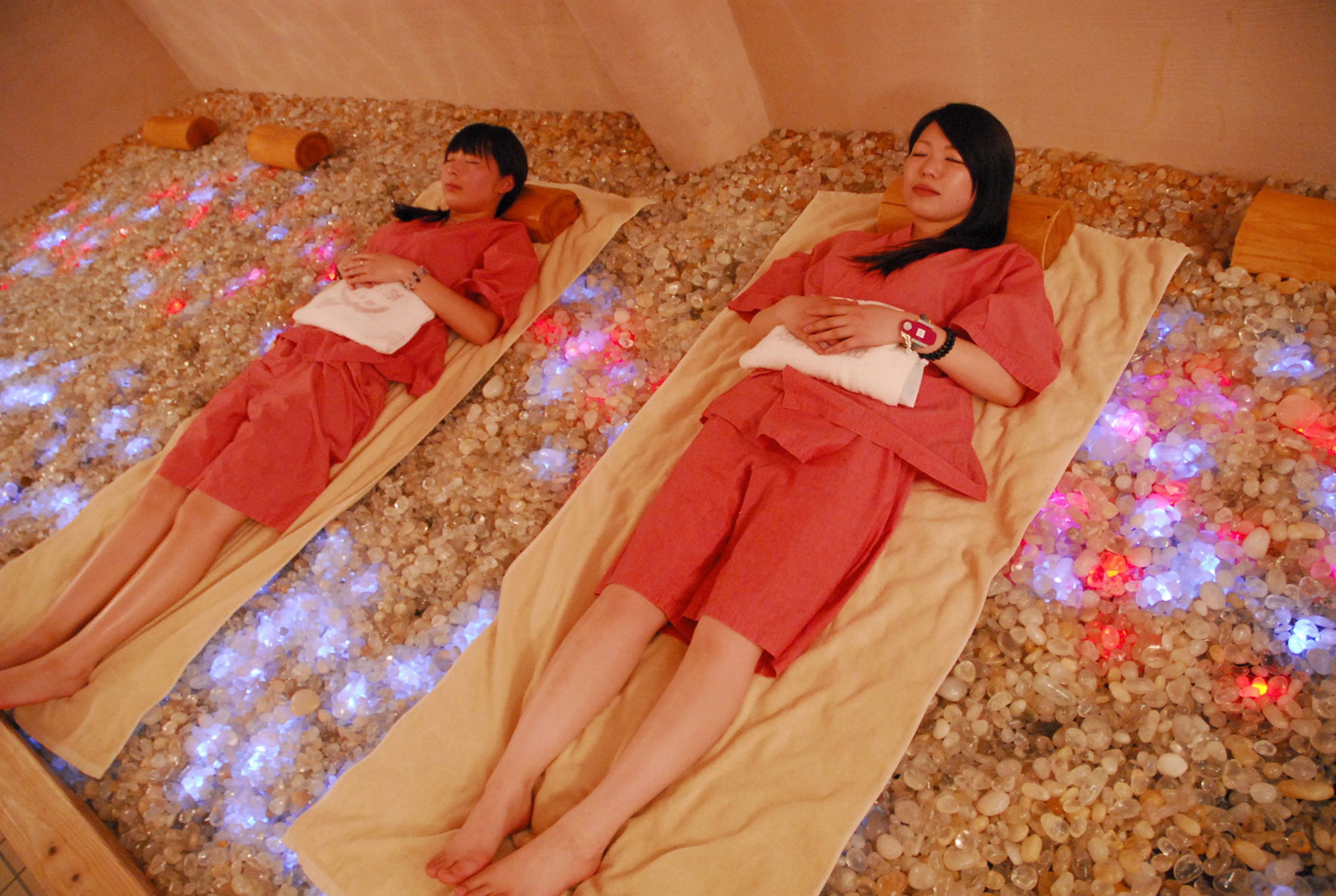 Two individuals lying on separate beds in a room with a pebble floor, each covered with a beige towel.