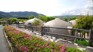 Una hilera de edificios con forma de cúpula, un paseo pavimentado y una valla de madera, rodeado de vegetación y montañas al fondo.