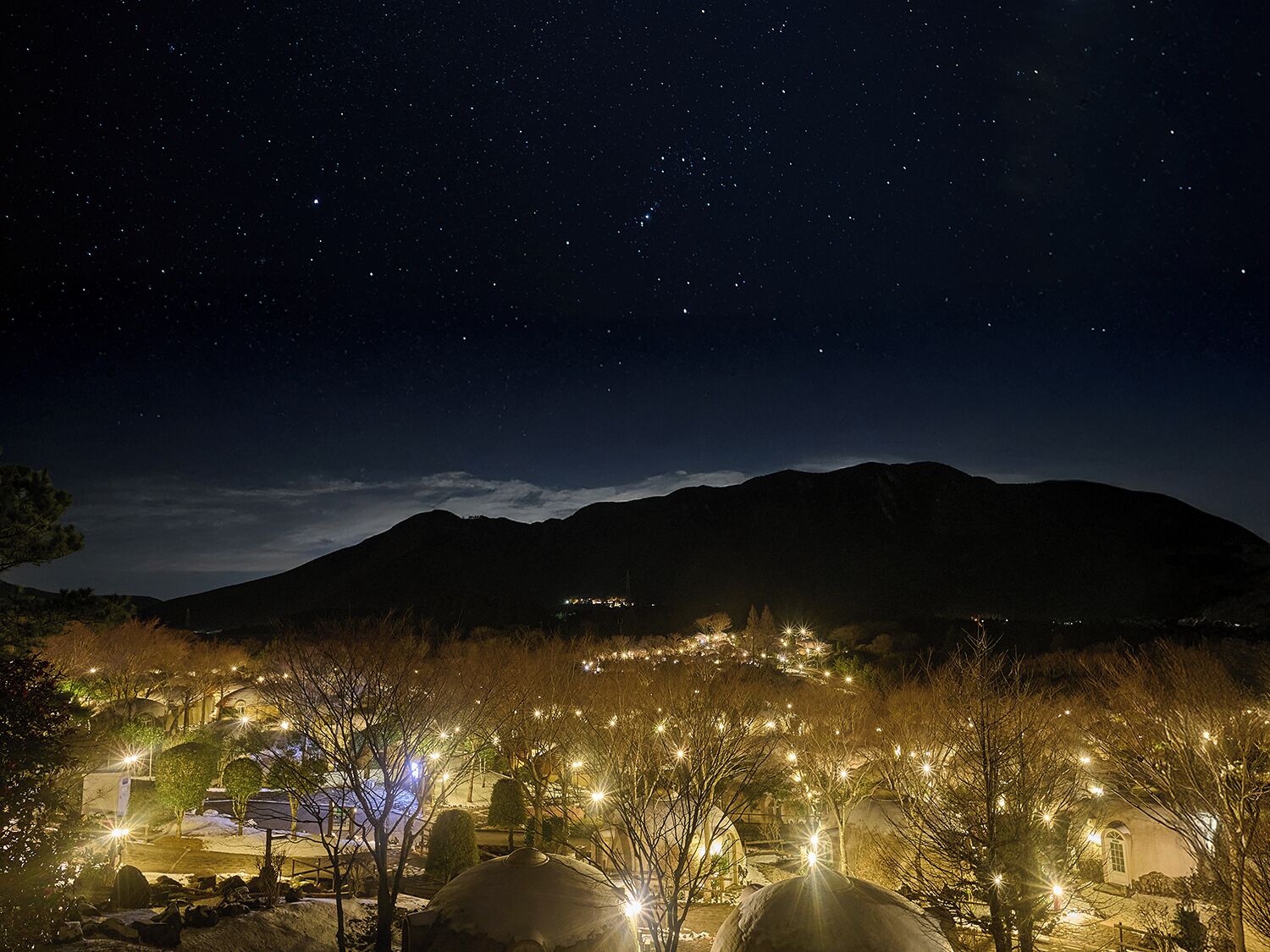 Vista nocturna de un pueblo con una montaña al fondo, edificios iluminados y un cielo estrellado.