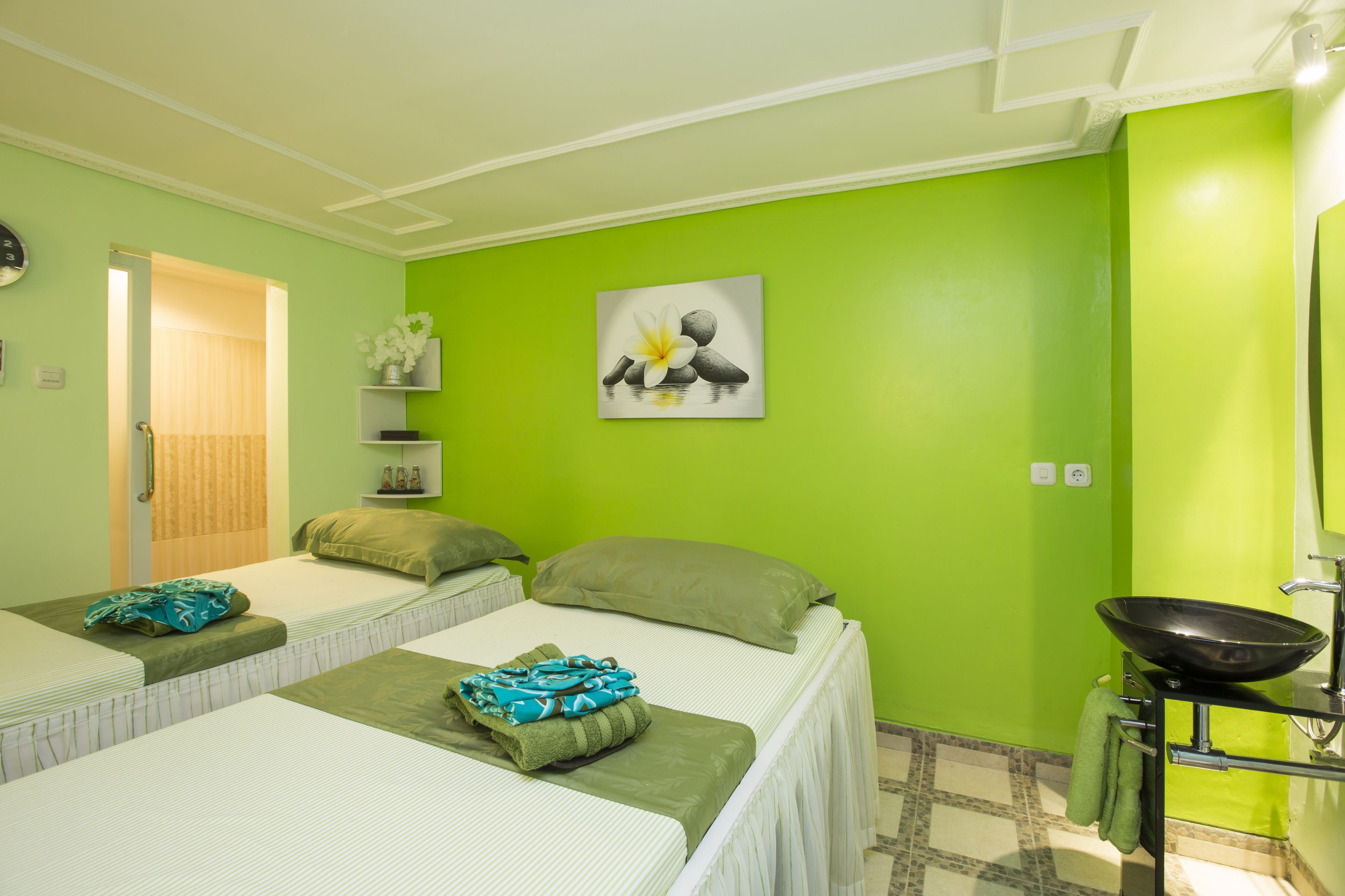 A room with two beds, a green wall, a painting, a sink, and a door leading to another room.