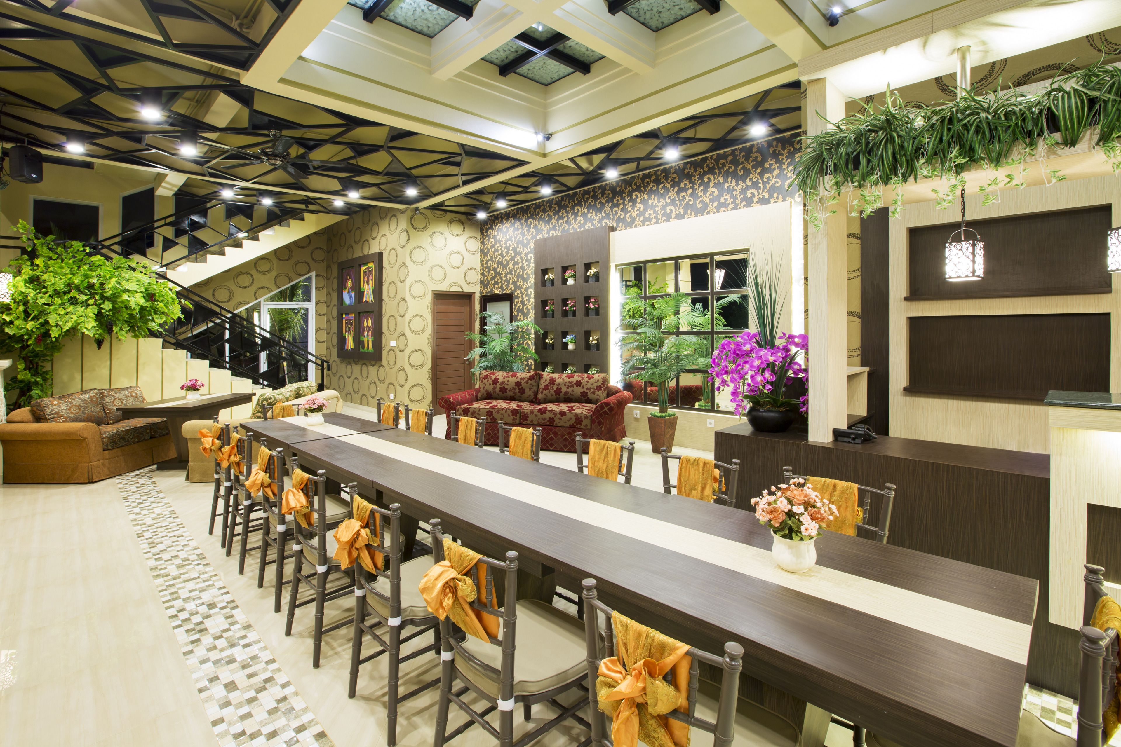 A spacious dining area with a long table, chairs with orange sashes, a staircase, and a sofa.