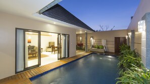 Un área de piscina exterior moderna con un ventanal corredera que da a una sala de estar, un pequeño rincón de descanso con cojines y una terraza de madera alrededor de la piscina.
