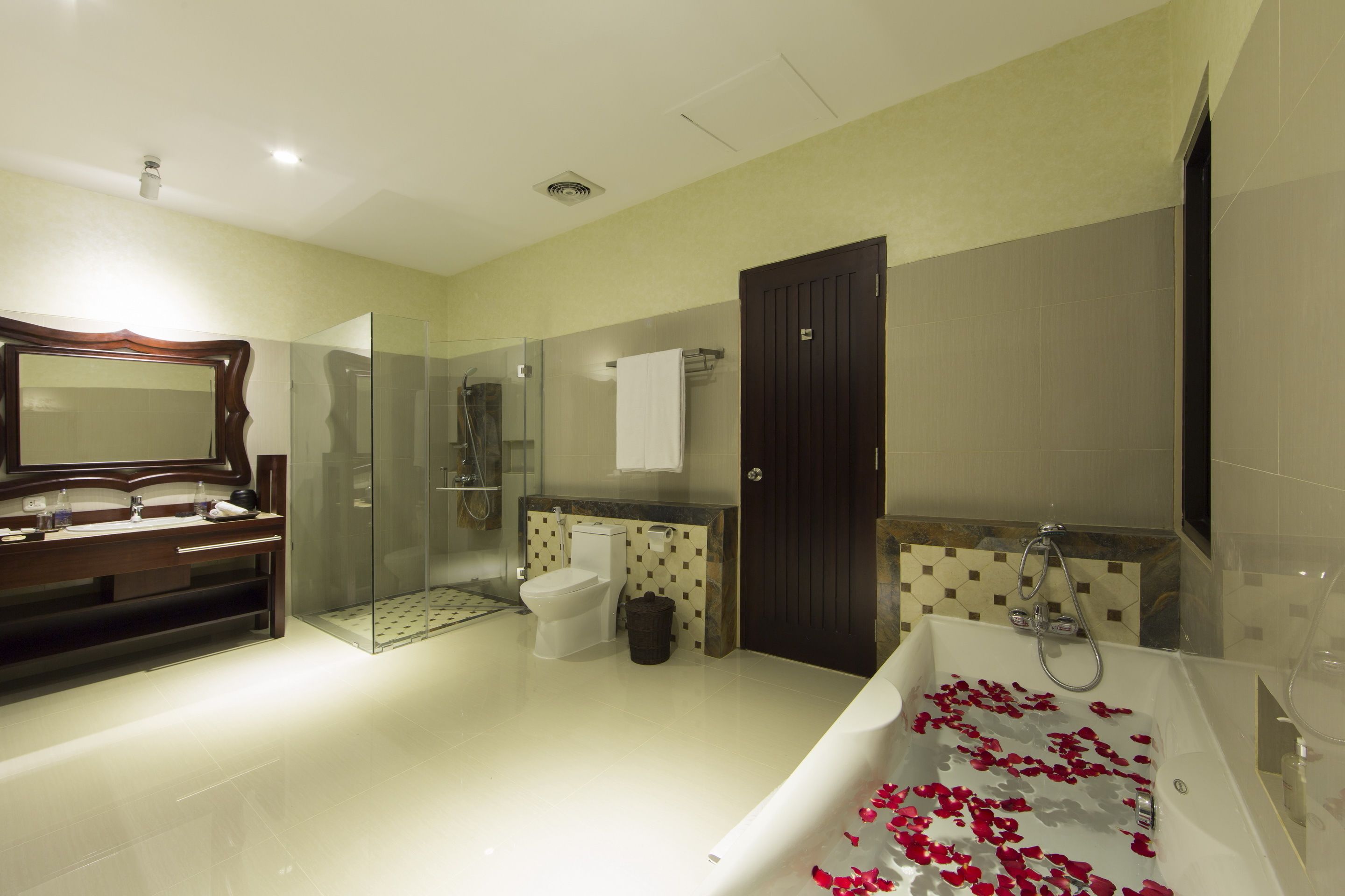 Une salle de bain moderne avec une douche à l’italienne en verre, une baignoire blanche parsemée de pétales de roses, un meuble-lavabo en bois et un grand miroir.