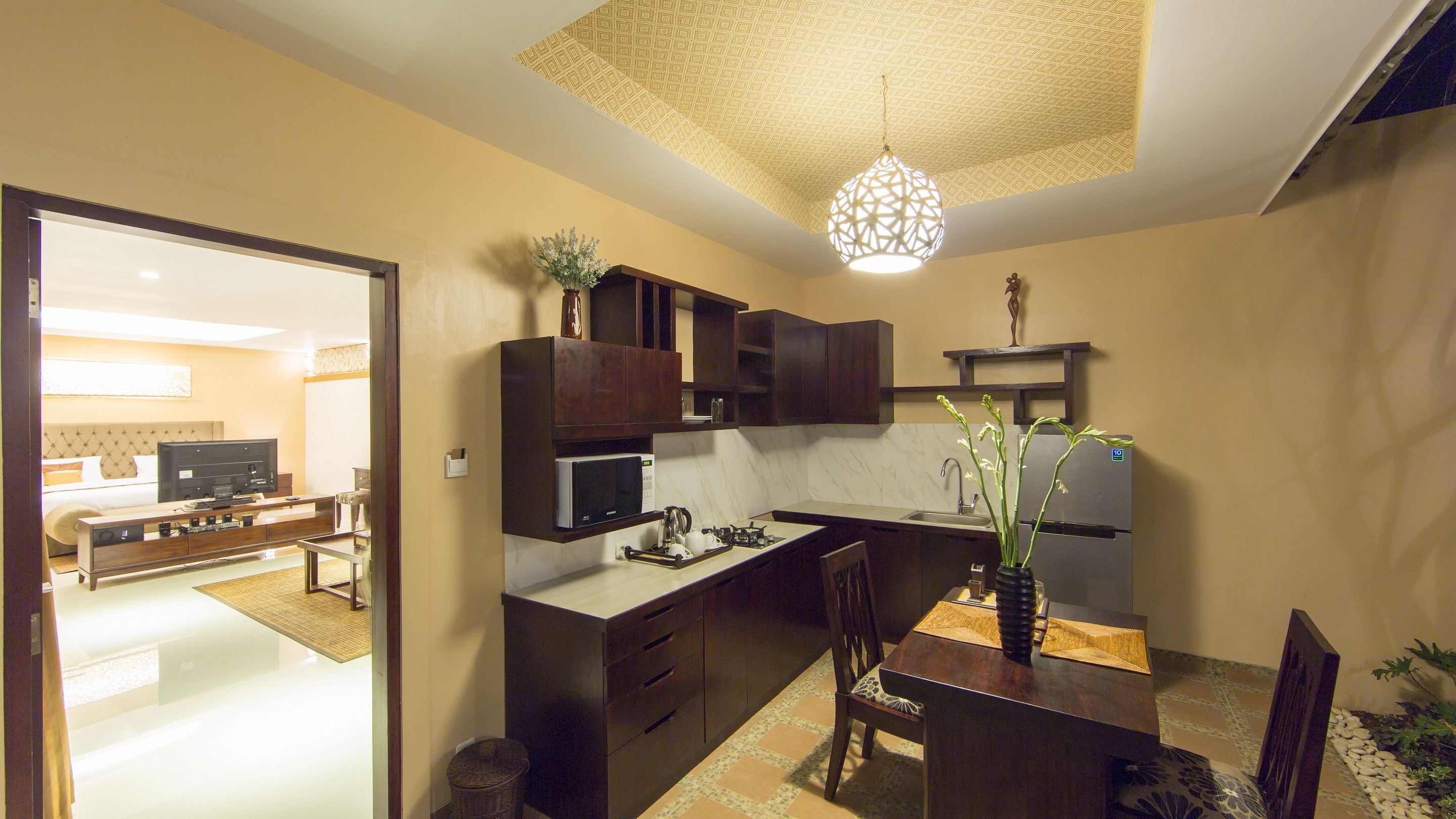 A compact kitchen with dark wood cabinets, a microwave, a gas stove, and a refrigerator. There's a dining table with chairs and a vase of flowers.